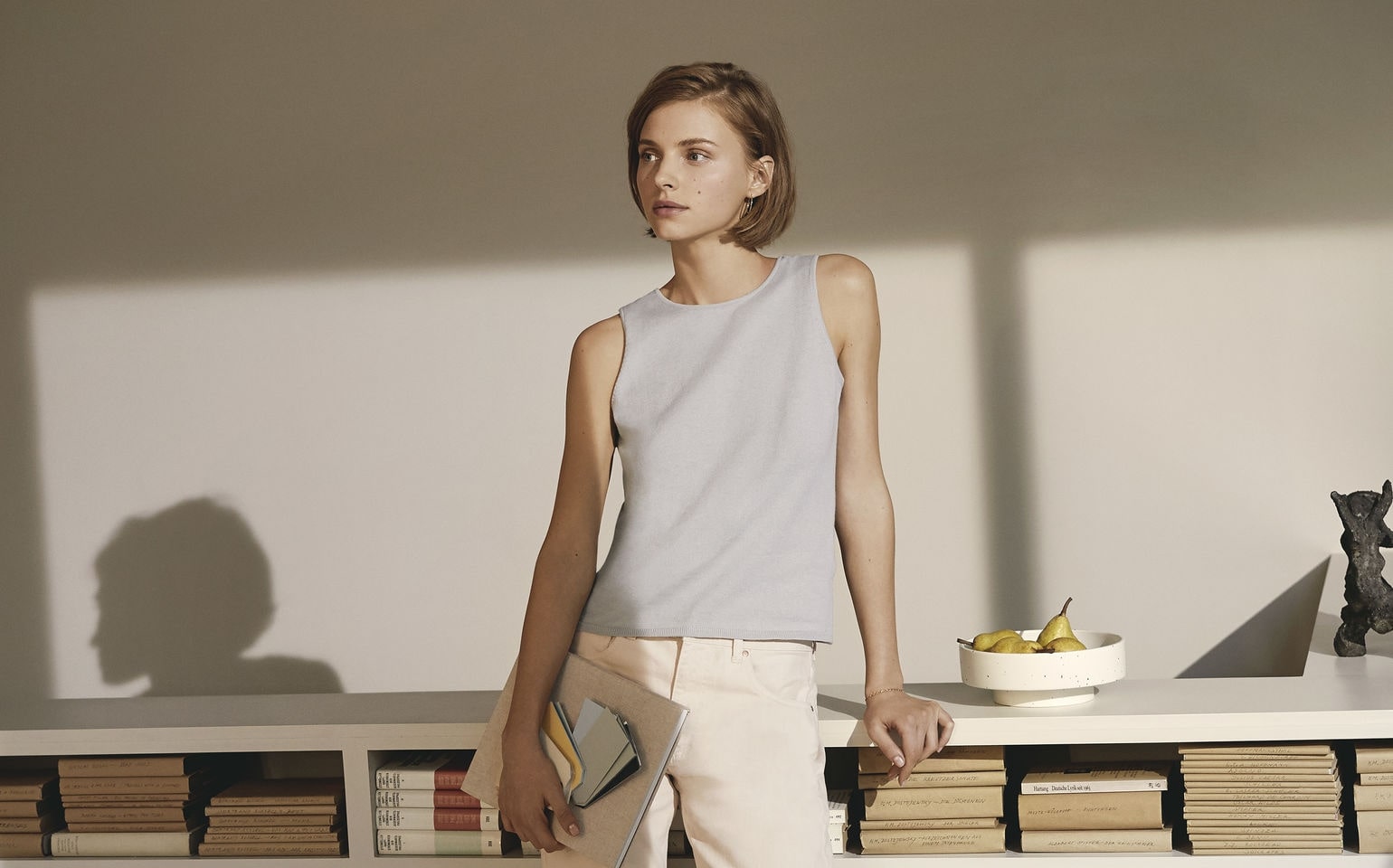 A casually dressed woman stands by a bookshelf, holding a book, in a calm, softly lit setting with neutral tones.